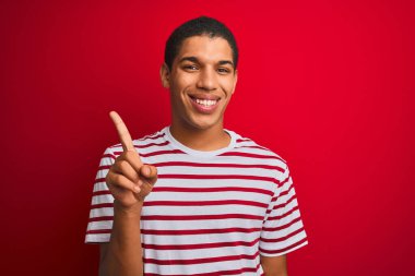 İzole edilmiş kırmızı arka plan üzerinde çizgili tişört giyen genç yakışıklı Arap adam bir numaralı parmağını gösterirken kendinden emin ve mutlu bir şekilde gülümsüyor..