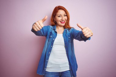 İzole edilmiş pembe arka plan üzerinde duran güzel, kızıl saçlı kadın el hareketlerini onaylıyor, gülümsüyor ve başarı için mutlu. Kazanan jest.