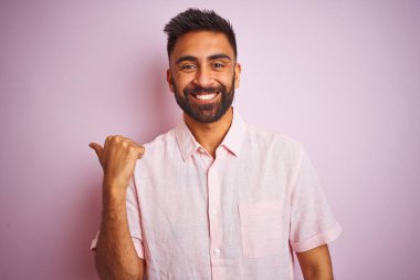 Izole pembe arka plan üzerinde duran rahat gömlek giyen genç hintli adam mutlu yüz görünümlü gülümseyerek ve başparmak ile yan işaret.