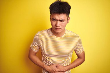 Asyalı genç bir Çinli tişört giyiyor. İzole edilmiş sarı arka planda duruyor. Midesinde mide bulantısı var. Ache kavramı.