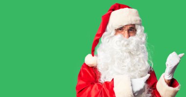 Orta yaşlı yakışıklı adam, Noel Baba kostümü ve sakal takmış. Sırtını işaret ediyor. El ve baş parmakları havada, kendine güvenen.