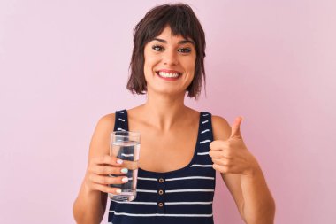 Genç, güzel bir kadın, pembe arka planda duran su bardağını tutuyor. Kocaman gülümsüyor ve gülümsüyor. Tamam işareti yapıyor.