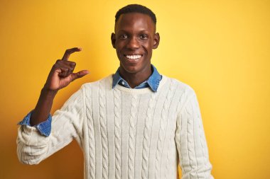 İzole edilmiş sarı arka plan üzerinde kot gömlek ve beyaz kazak giyen Afro-Amerikan adam gülümsüyor ve el hareketleriyle küçük boy işareti yaparak kameraya bakıyor. Ölçü kavramı.