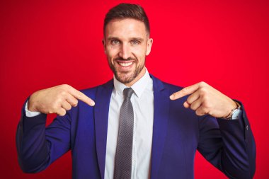 Genç ve yakışıklı bir iş adamı, izole edilmiş kırmızı bir arka planda, kendine güvenen, yüzünde bir gülümsemeyle, kendini gururlu ve mutlu olarak gösteriyor..