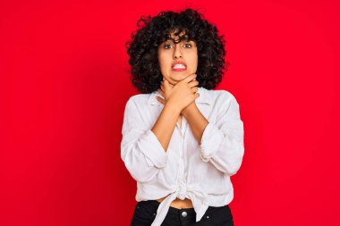 Young arab woman with curly hair wearing white casual shirt over isolated red background shouting and suffocate because painful strangle. Health problem. Asphyxiate and suicide concept.