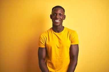 Sıradan tişörtler giyen genç Afro-Amerikalı adam izole edilmiş sarı arka planda el ele tutuşmuş ve mutlu bir şekilde gülümsüyor. Başarı ve iyimser
