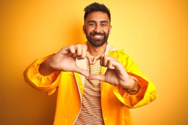 İzole edilmiş sarı arka planda duran yağmurluk giyen genç Hintli adam elleriyle kalp sembolü yapan aşka gülümsüyor. Romantik konsept.