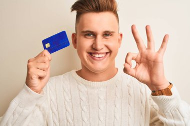 Genç ve yakışıklı bir adam kredi kartını soyutlanmış beyaz arka planda tutuyor. Parmaklarıyla imza atıyor. Mükemmel bir sembol.