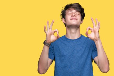 İzole arka plan üzerinde mavi t-shirt giyen genç yakışıklı adam rahatla ve gülümseyen gözlerle kapalı parmak jestle meditasyon yapıyor. Yoga kavramı.