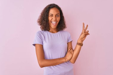 İzole edilmiş pembe arka planda tişört giyen genç Brezilyalı kadın kameraya gülümseyen ve parmaklarıyla zafer işareti yapan. İki numara..