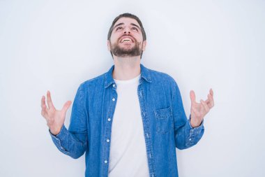 Beyaz izole edilmiş arka planda kot ceket giyen genç, yakışıklı bir İspanyol çılgın ve sinirli bir şekilde bağırıyor ve agresif bir ifade ve kollarla bağırıyor. Hayal kırıklığı konsepti.