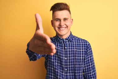 İzole edilmiş sarı arka plan üzerinde duran sıradan tişörtlü genç yakışıklı adam dostça bir selamlama ve hoş geldin hediyesi olarak el sıkışma sunuyor. Başarılı bir iş..