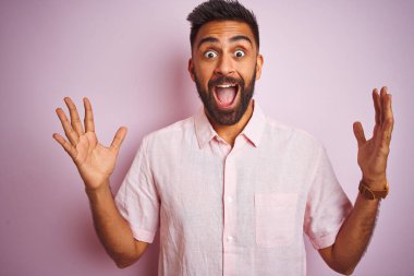 İzole edilmiş pembe arka plan üzerinde duran genç Hintli adam çılgın ve başarıyı kollarını kaldırıp heyecanla haykırarak kutluyor. Kazanan konsept