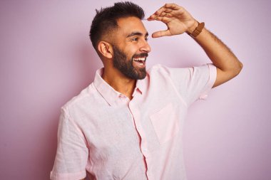 İzole edilmiş pembe arka planda dikilen rahat gömlekli genç Hintli adam çok mutlu ve tebessüm ediyor. Konsept aranıyor.