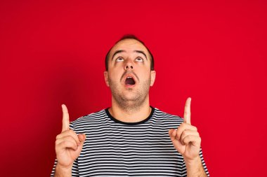 İzole edilmiş kırmızı arka plan üzerinde duran çizgili donanma tişörtü giyen genç adam şaşırmış ve şaşırmıştı. Parmaklarıyla işaret ediyor ve kollarını kaldırıyordu..