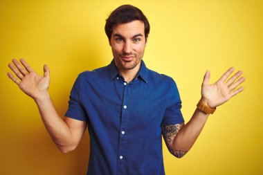 Üstü açık tişört giyen, dövmeli genç bir adam izole edilmiş sarı arka planda duruyor. Hiçbir fikri yok ve elleri ve kolları havada şaşkın bir ifadesi var. Şüphe kavramı.