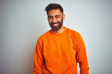 İzole edilmiş beyaz arka plan üzerine turuncu kazak giyen genç Hintli adam kameraya seksi bir ifadeyle bakıyor, neşeli ve mutlu bir yüzle..
