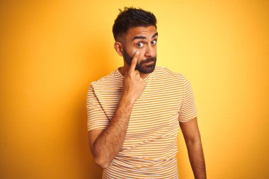İzole edilmiş sarı arka planda duran tişörtlü genç Hintli adam senin jestini, şüpheli ifadesini izliyor.
