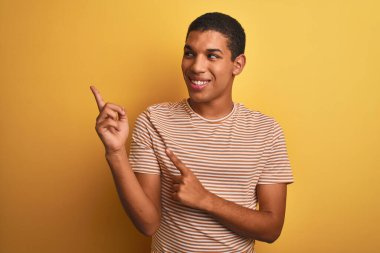 Çizgili tişört giyen yakışıklı Arap adam izole edilmiş sarı arka planda duruyor gülümsüyor ve kameraya bakıyor iki eliyle ve parmaklarıyla yan tarafa bakıyor..