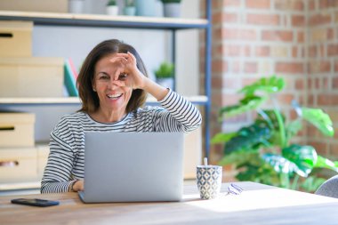 Orta yaş yaşlı kadın evde masada oturan mutlu yüzü ile bilgisayar dizüstü bilgisayar kullanarak çalışan parmakları ile bakarak göz üzerinde el ile ok işareti yapıyor gülümseyerek