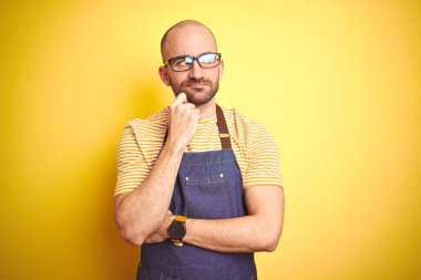 Kahveci önlüğü giymiş genç bir barmen sarı arka planda çalışırken elleri çenesinde soru sorarken, düşünceli bir ifade takınıyor. Düşünceli bir yüzle gülümsüyor. Şüphe kavramı.