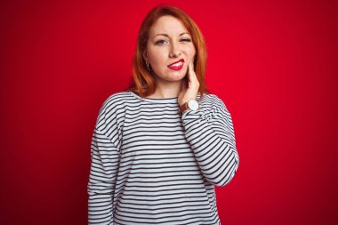 Kızıl saçlı genç bir kadın donanma gömleği giyiyor kırmızı izole arka planda duruyor diş ağrısı ya da diş hastalığından dolayı acı dolu bir ifadeyle ağıza dokunuyor. Dişçi konsepti.