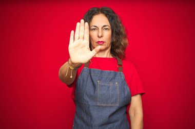 Kırmızı izole arka planda önlük giyen orta yaşlı bir kadın avuç içiyle şarkı söylemeyi kesiyor. Yüzünde olumsuz ve ciddi bir jest olan uyarı ifadesi.