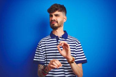 İzole edilmiş mavi arka planda duran çizgili polo giyen genç adam tiksindirici ve tiksindirici bir ifade takınıyor çünkü tiksinti tepki veriyor. Eller havada. Can sıkıcı bir kavram..
