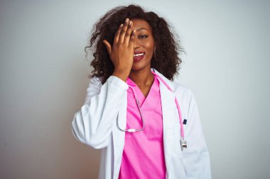 İzole edilmiş beyaz arka plan üzerine pembe steteskop takmış Afro-Amerikalı bir kadın. Bir gözünü elinle kapatıyor, yüzünde kendine güvenen bir gülümseme ve sürpriz bir duygu var..