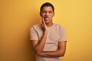 İzole edilmiş sarı arka plan üzerinde duran çizgili tişört giyen genç, yakışıklı Arap adam diş ağrısı ya da diş hastalığından dolayı acı dolu bir ifadeyle ağıza dokunuyor. Dişçi konsepti.