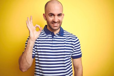 Sakallı, sarı izole edilmiş arka plan üzerine sıradan mavi tişörtlü genç, kel bir adam gülümseyerek el ve parmaklarıyla tamam işareti yapıyor. Başarılı ifade.