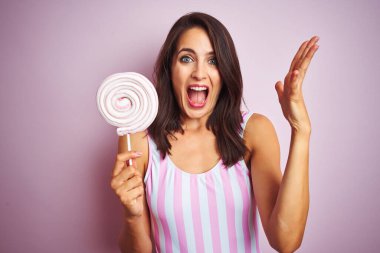 Genç güzel kadın pembe izole arka plan üzerinde tatlı şeker yeme çok mutlu ve heyecanlı, kazanan ifade zafer büyük gülümseme ile çığlık kutluyor ve el kaldırdı