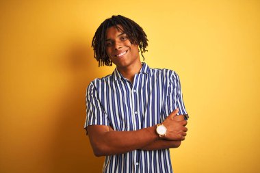 Rastgele çizgili tişört giyen Afro bir adam izole edilmiş sarı arka plan üzerinde kollarını kavuşturup kameraya bakarken gülümseyen mutlu bir yüz. Pozitif kişi.