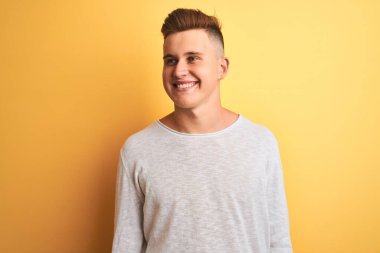 İzole edilmiş sarı arka plan üzerinde duran beyaz tişört giyen yakışıklı genç adam yüzünde gülümseme, doğal bir ifade ile yan yana bakıyor. Kendinden emin gülen.