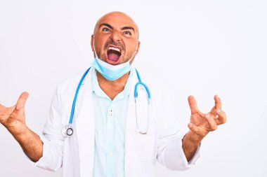 Orta yaşlı doktor steteskop takıyor ve maske takıyor beyaz arka planda deli deli deli bağırıyor ve bağırıyor agresif bir ifade ve kollar havada. Hayal kırıklığı konsepti.