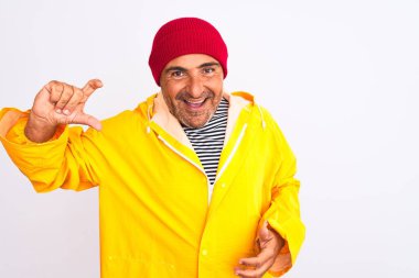 İzole edilmiş beyaz arka planda duran yünlü şapkalı ve yağmurluk giyen orta yaşlı bir adam gülümsüyor ve el hareketleriyle küçük boy işareti yapıyor ve kameraya bakıyor. Ölçü kavramı.