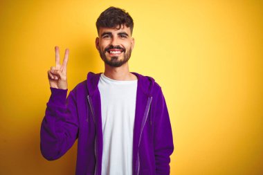 İzole edilmiş sarı arka plan üzerine mor kazak giyen dövmeli genç adam iki numaralı parmaklarıyla gülümseyerek kendinden emin ve mutlu görünüyor..