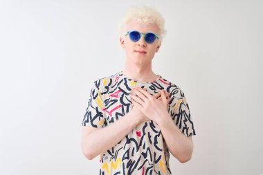 Renkli tişört ve güneş gözlüğü takan genç albino adam izole edilmiş kırmızı arkaplanda elleri göğsünde, gözleri kapalı ve yüzünde minnet dolu bir jest ile gülümsüyor. Sağlık kavramı.