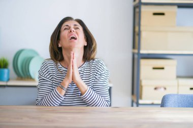 Orta yaş yaşlı kadın evde masada oturan dilencilik ve umut ifadesi ile birlikte elleri ile dua çok duygusal ve endişeli yüzünde. Af dilemek istiyorum. Din kavramı.