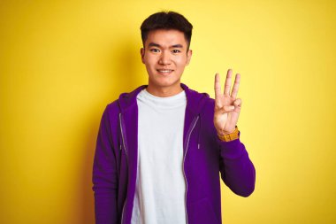 Mor kazak giyen Asyalı genç adam izole edilmiş sarı arka planda duruyor ve üç numaralı parmaklarıyla işaret ediyor kendinden emin ve mutlu gülümserken..