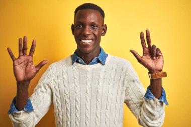 İzole edilmiş sarı arka plan üzerinde kot gömlek ve beyaz kazak giyen Afro-Amerikalı adam sekiz numaralı parmaklarıyla işaret ederken kendinden emin ve mutlu bir şekilde gülümsüyor..
