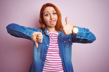 Güzel kızıl saçlı kadın kot gömlek ve çizgili tişört giyerek pembe arka planda baş parmak hareketi, anlaşmazlık ve uzlaşma ifadesi yapıyor. Çılgın çatışma