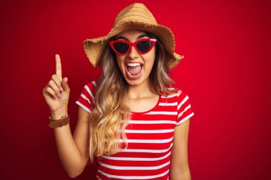 Güneş gözlüğü takan genç ve güzel bir kadın ve kırmızı izole edilmiş arka plan üzerine yaz şapkası takmış başarılı bir fikir işaret ediyor. Heyecanlı ve mutlu. Bir numara..