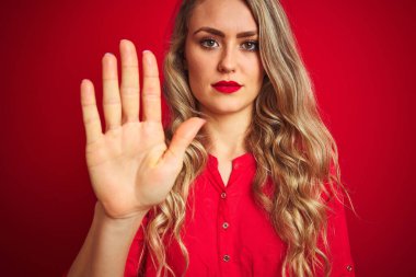 Güzel bir kadın kırmızı izole edilmiş arka planda duruyor, eli açık, dur işareti yapıyor, ciddi ve kendinden emin bir ifadeyle, savunma hareketi yapıyor.