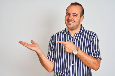 İzole edilmiş beyaz arka planın üzerinde duran mavi çizgili genç adam hayrete düşmüş ve kameraya gülümsüyor..