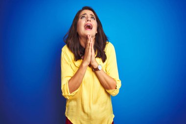 Sarı tişörtlü genç bir kadın mavi izole edilmiş arka planda duruyor. Elleri bağlı yalvarıyor ve dua ediyor. Yüzünde umut ifadesi var. Af diliyor. Din kavramı.