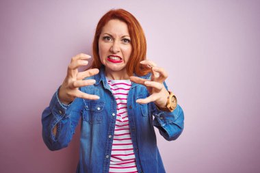 Güzel kızıl saçlı kadın kot gömlek ve çizgili tişört giyip izole edilmiş pembe arka planda öfkeyle haykırıyor, elleri boğmaya çalışıyor, deli gibi bağırıyor.