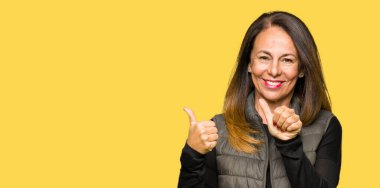 Orta yaş kadın giyiyor kış yelek işaretleme için arkasından el ve başparmak yukarıya, kendinden emin gülümseyen güzel