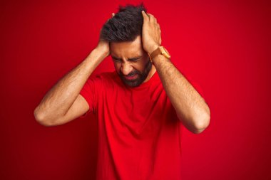 Genç, yakışıklı Hintli adam, izole edilmiş kırmızı arka plan üzerine tişört giyip acı ve migren yüzünden çaresiz ve stresli bir baş ağrısı çekiyor. Ellerini başının üstüne koy..