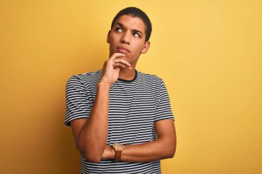 Genç yakışıklı Arap adam, izole edilmiş sarı arka plan üzerinde donanma çizgili tişört giyiyor ve elleri çenesinde soru sormayı, düşünceli ifadeyi düşünüyor. Düşünceli bir yüzle gülümsüyor. Şüphe kavramı.